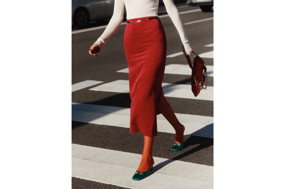 Fashionable woman strides across zebra crossing wearing red skirt, white top, green heels, carrying matching red handbag in daylight.
