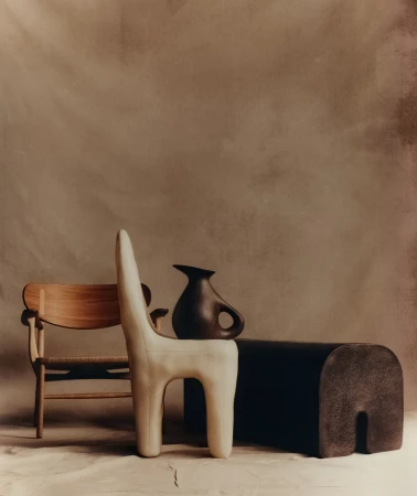 Minimalist studio still life with sculptural cream chair, dark ceramic jug, wooden chair, and low dark bench against textured backdrop