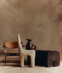 Minimalist studio still life with sculptural cream chair, dark ceramic jug, wooden chair, and low dark bench against textured backdrop
