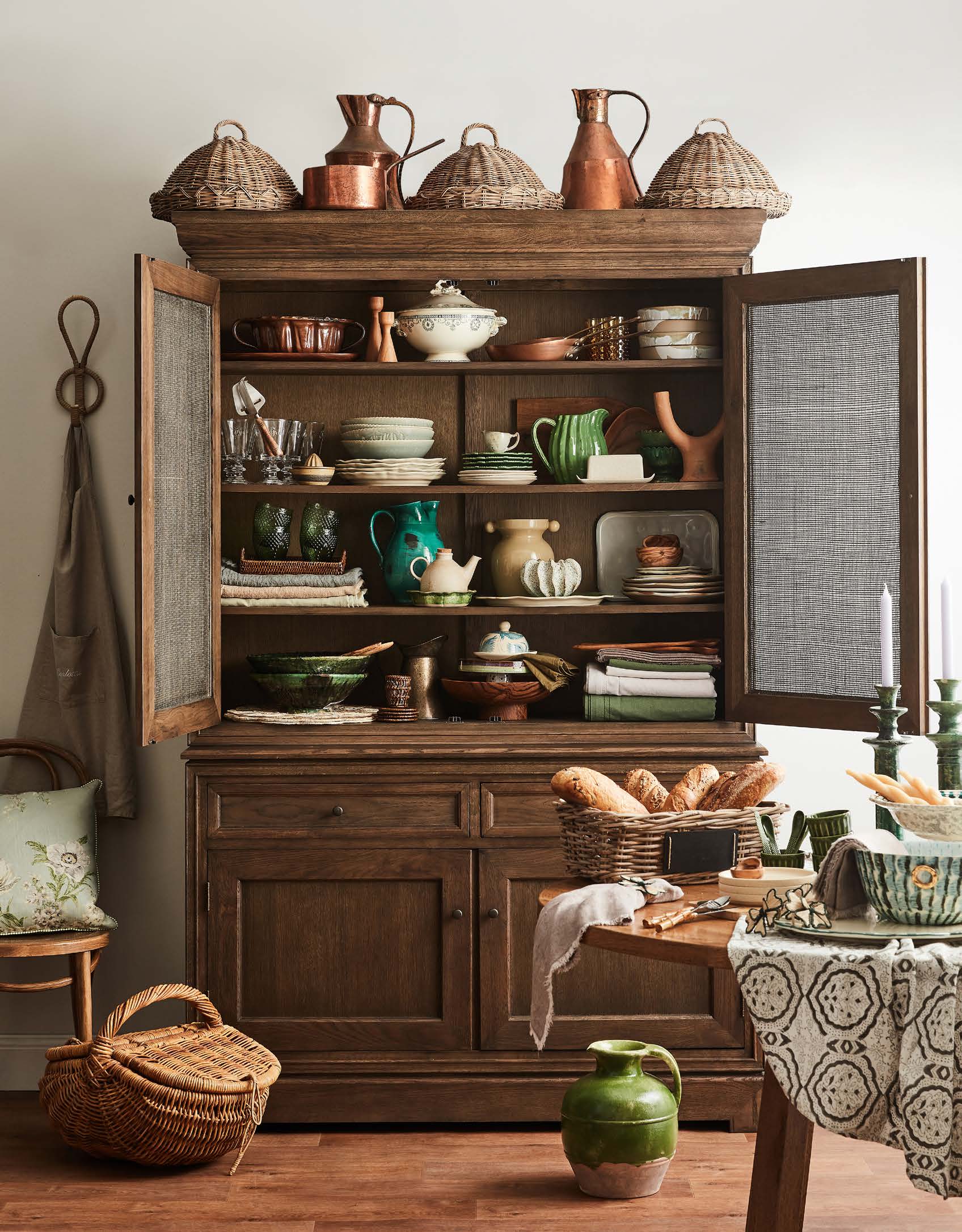 A wood hutch displays a collection of dishware, pottery, and copper pots in warm, inviting light, with a dining table nearby.