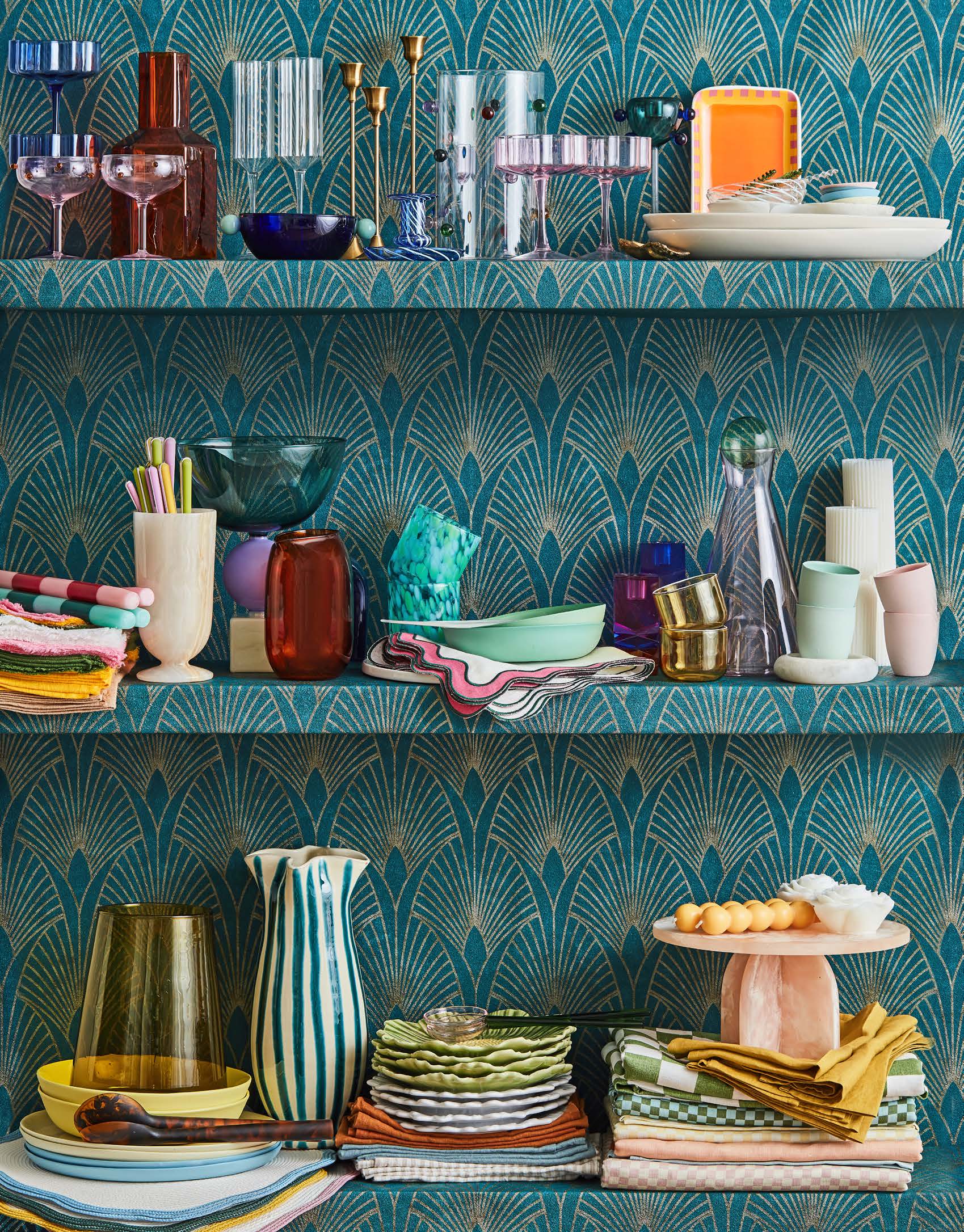 Still life photograph features an array of glassware and dishes displayed on a three-shelf unit against teal, patterned wallpaper.