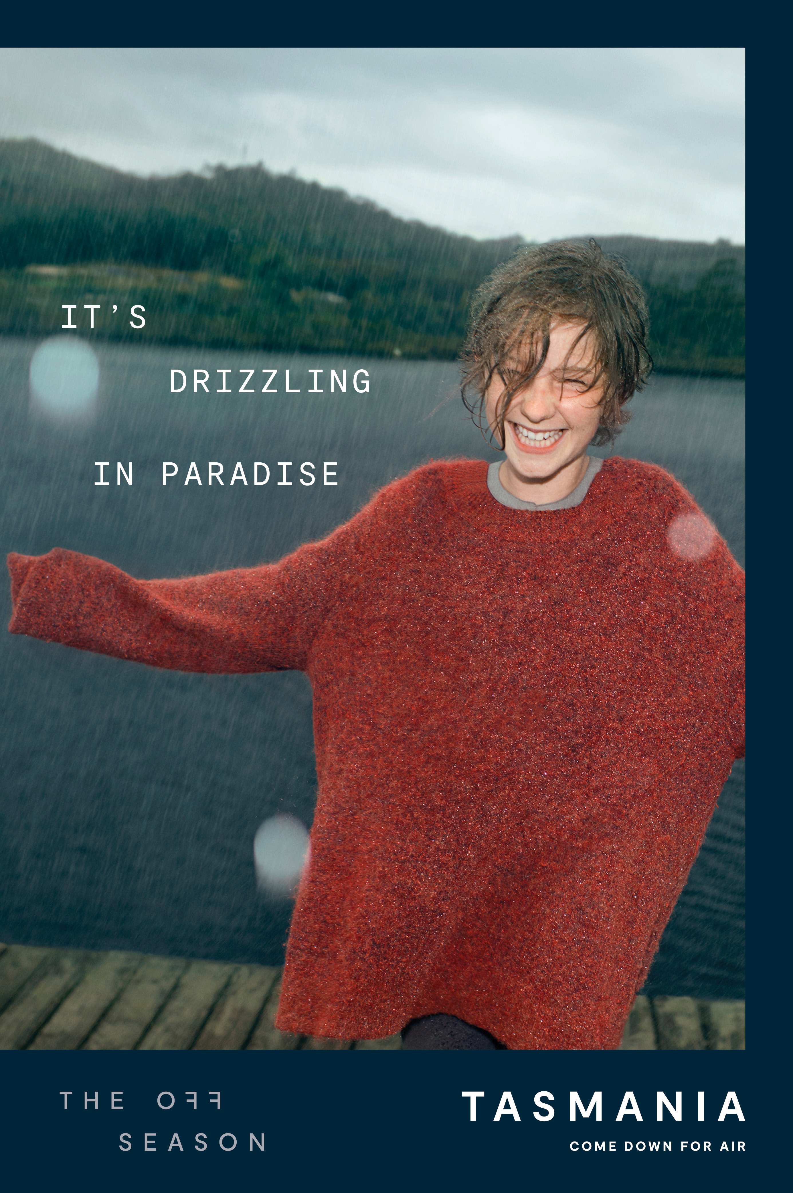 Young woman in oversized sweater smiling in the rain near a lake in Tasmania. Tourism ad with the text 'It's drizzling in paradise'.
