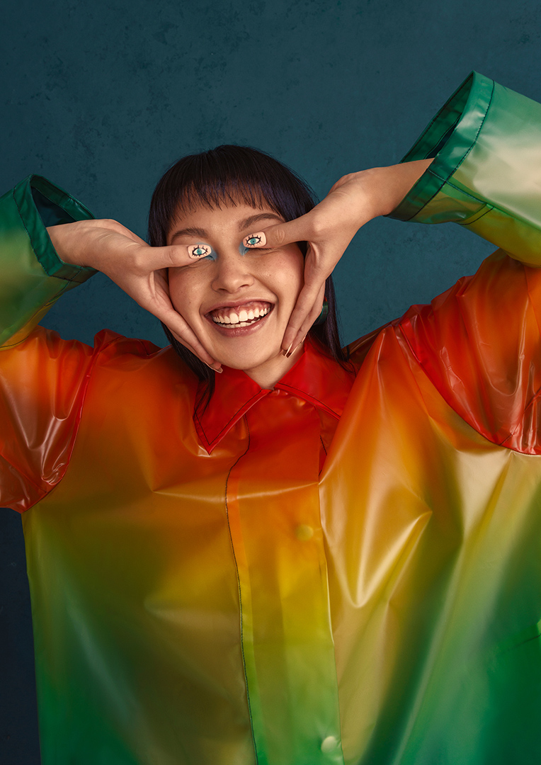 Smiling woman with dark hair and colorful transparent coat poses indoors with hand gestures near her face, cheerful mood.