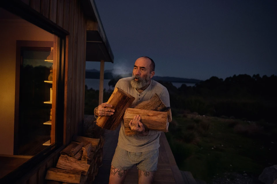Man carrying firewood outside a cabin at dusk. He is wearing shorts and a t-shirt near a lake and forest landscape at night.