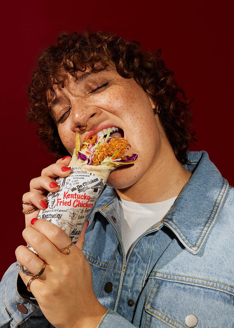 Woman with curly hair enjoying a chicken wrap in front of a maroon background, wearing a denim jacket and multiple rings. 