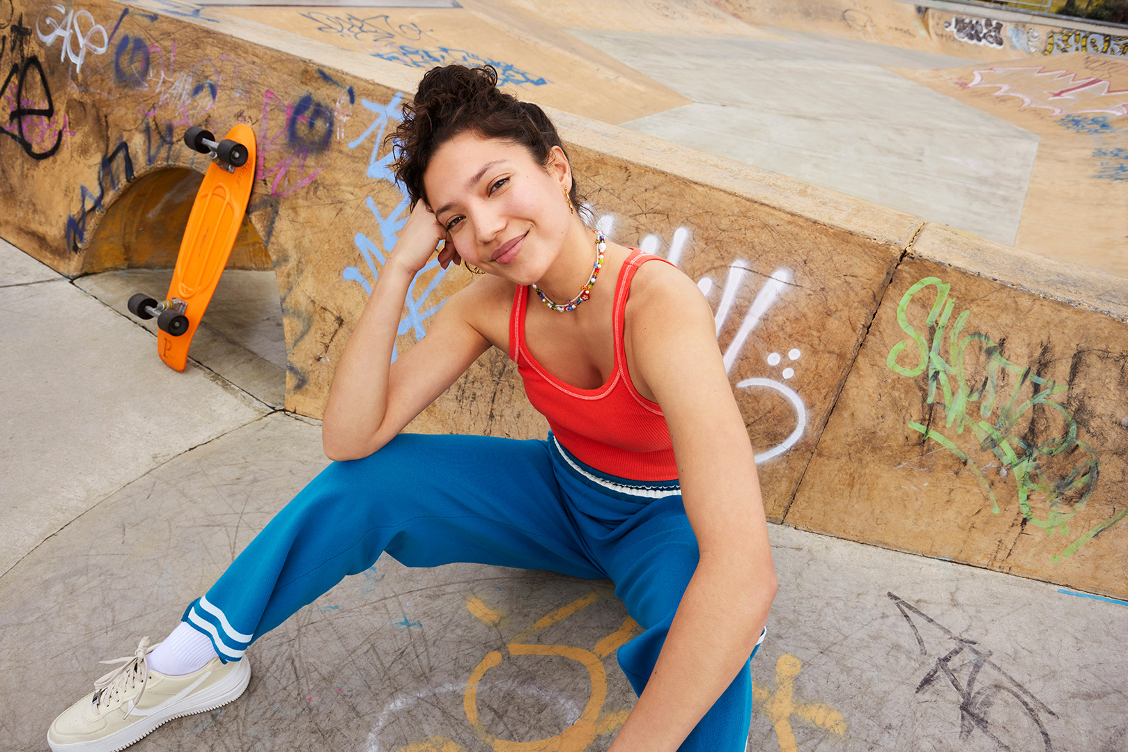 Young woman poses in blue sweatpants and a red top at a skatepark, sitting near a bright orange skateboard against graffiti walls.