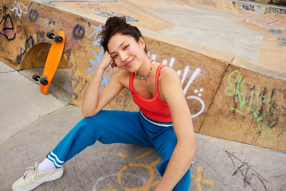 Young woman poses in blue sweatpants and a red top at a skatepark, sitting near a bright orange skateboard against graffiti walls.