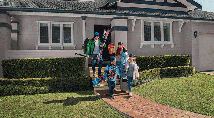 Family of four descends brick path from home, ready for winter vacation with skis, snowboards, luggage, and cold-weather attire.