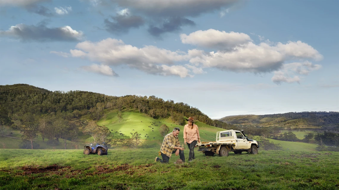 A couple works on a farm, kneeling on grassy land, with a truck and ATV nearby under a partly cloudy sky, showing an agricultural scene.
