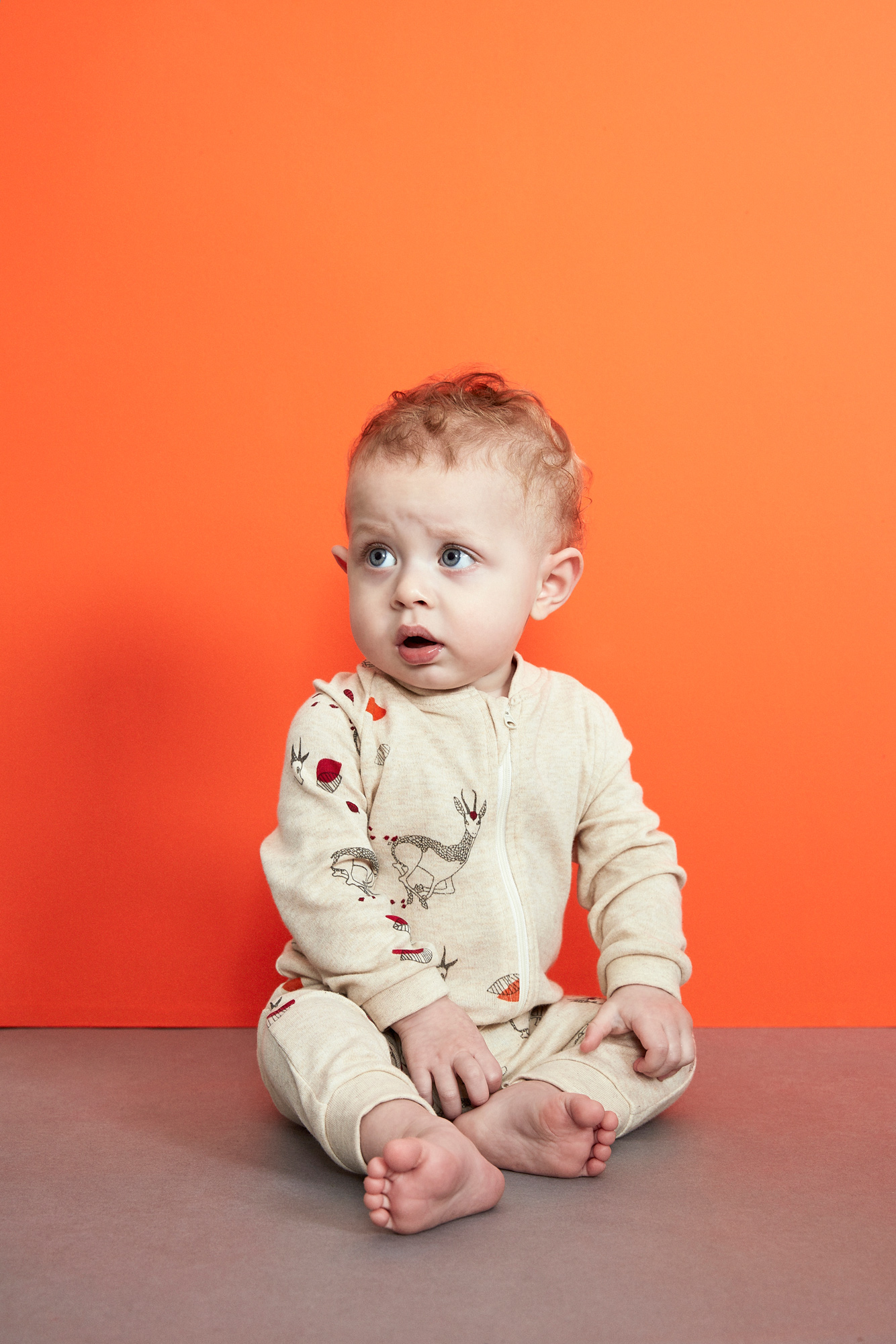 Adorable baby with curly hair sits on a gray floor against a bright orange background wearing a patterned onesie, looking surprised.