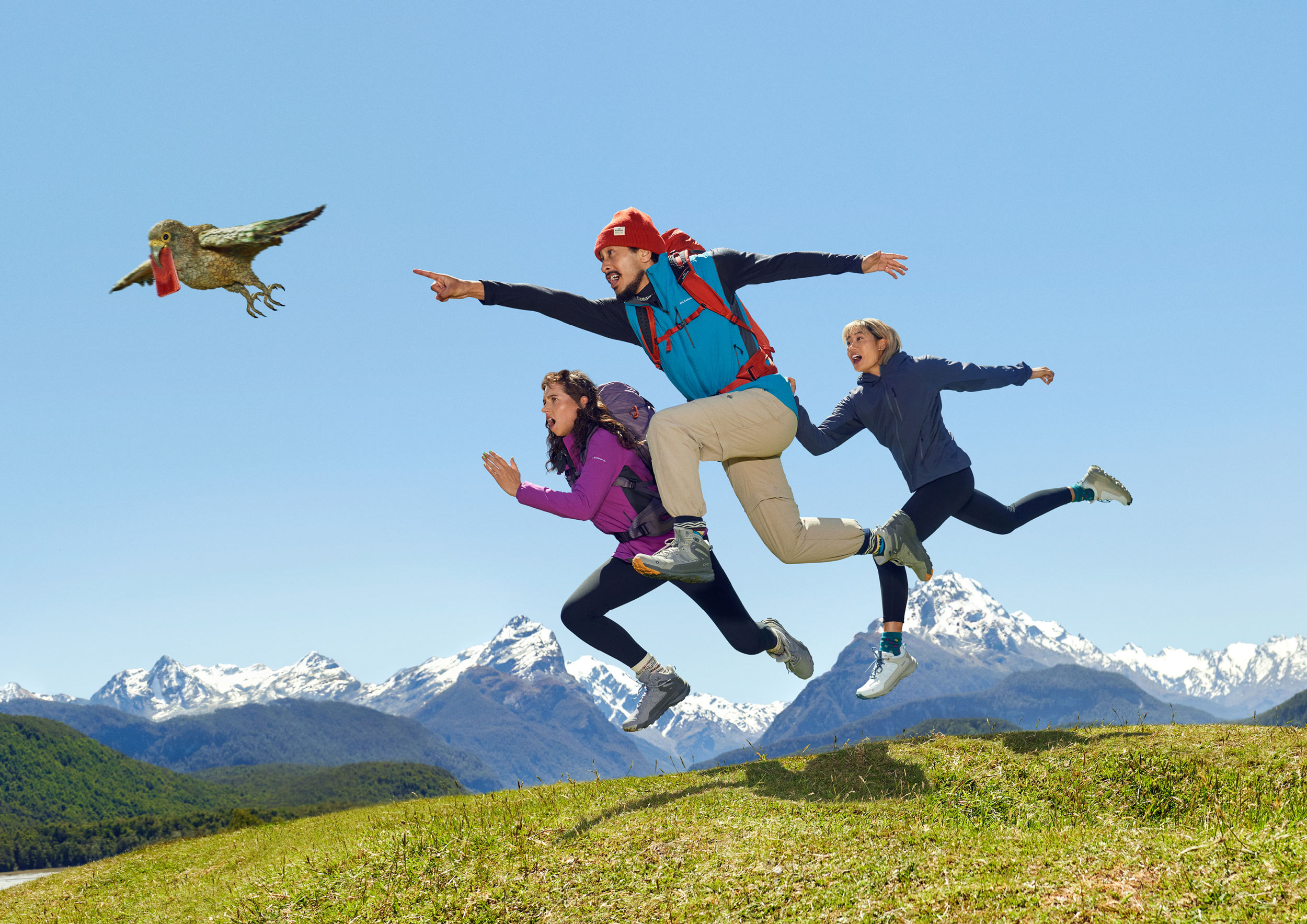 Three people in outdoor apparel jump in the air on a grassy hill with snow-capped mountains in the background as a bird flies by.