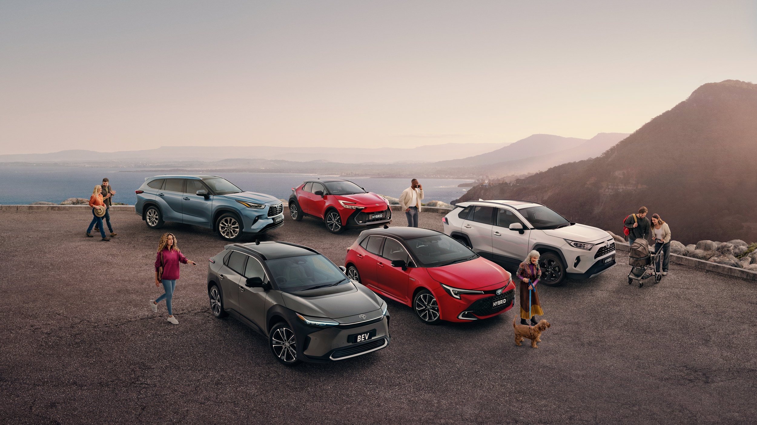 A variety of Toyota vehicles including the bZ4X EV, Corolla, and RAV4, are parked on a scenic overlook near mountains and ocean with groups of people.