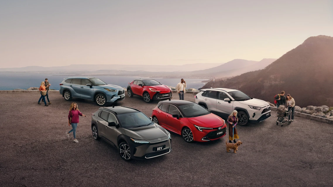 A variety of Toyota vehicles including the bZ4X EV, Corolla, and RAV4, are parked on a scenic overlook near mountains and ocean with groups of people.