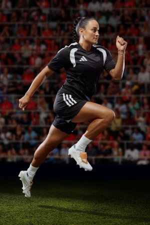 Action shot of female soccer player sprinting on field with blurred stadium background, wearing Adidas uniform and cleats.