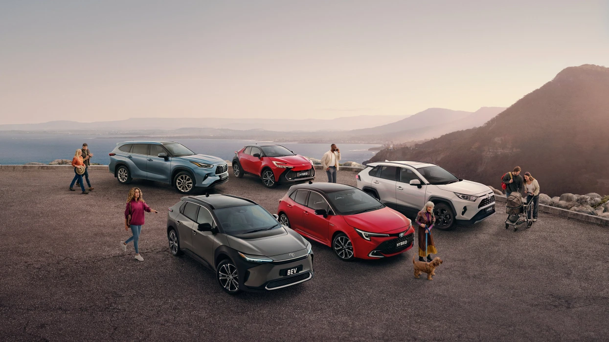 Multiple modern vehicles parked on a coastal overlook with people and a dog enjoying the scenic ocean view at sunset.