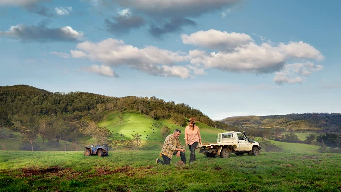 Two people working in a green pasture with a white utility truck and rolling hills under a cloudy sky