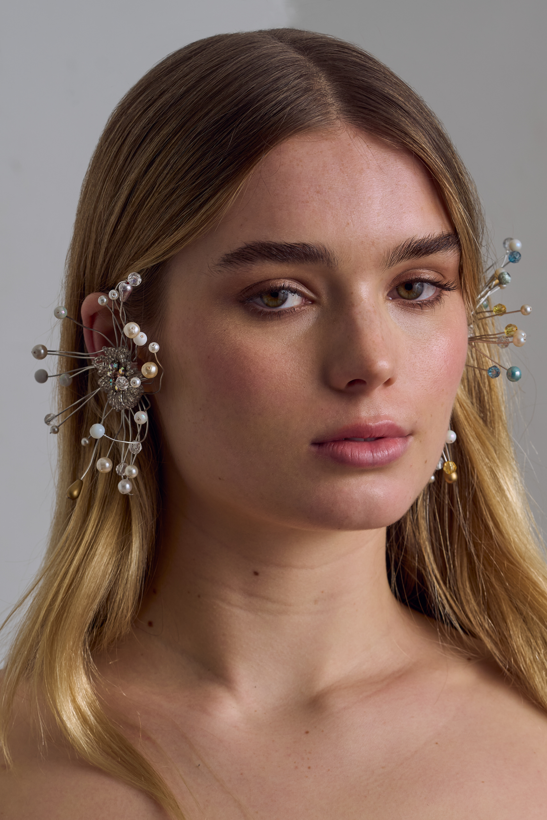 Close-up portrait of a fair-skinned woman with fair hair and elaborate ear jewelry against a neutral background in soft, diffused light.