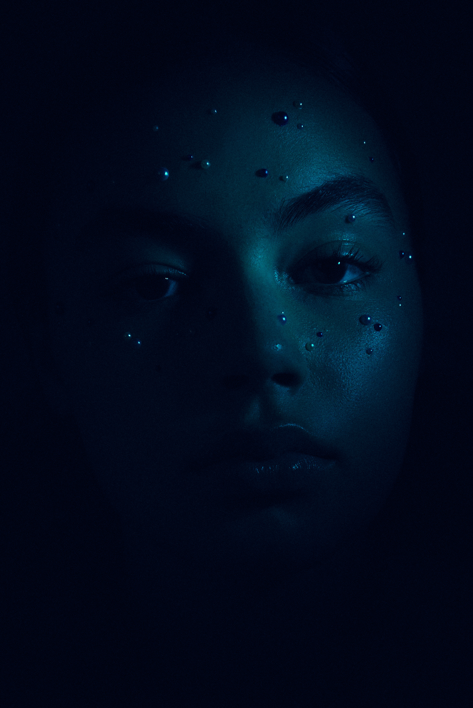 Close-up portrait of a woman's face in dark teal lighting with water droplets on her forehead and cheeks in a studio environment.