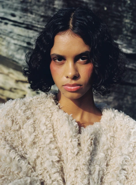 Portrait of a young adult woman with dark curly hair, wearing a fuzzy cream-colored jacket, against a blurred, textured rock background.