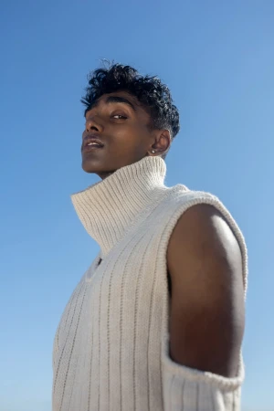 Low angle portrait of a man in a ribbed cream turtleneck sleeveless sweater against a clear blue sky background.