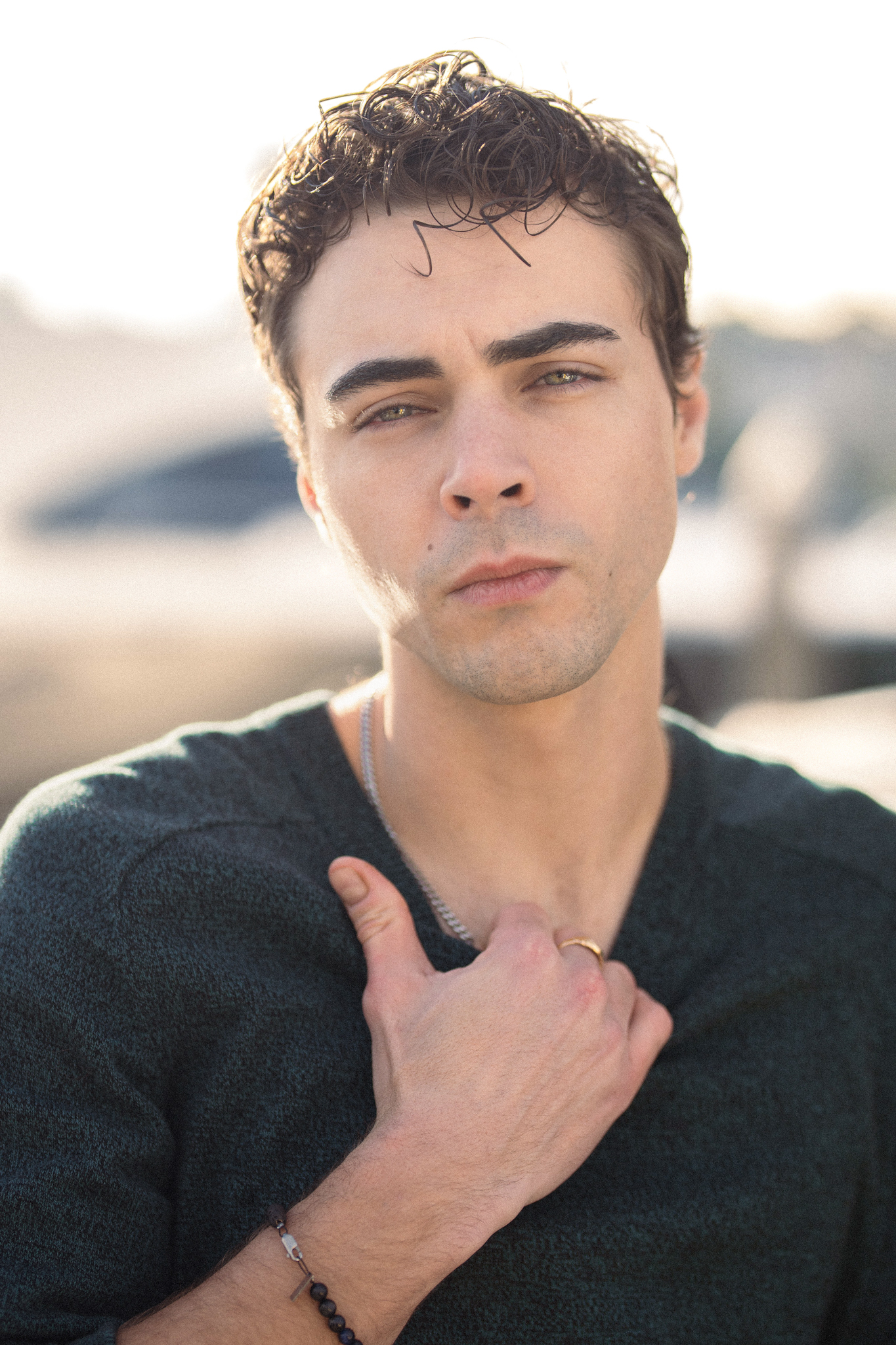 Portrait of a young adult male with dark hair, wearing a green sweater, posing outdoors in soft natural light, looking directly at the camera.