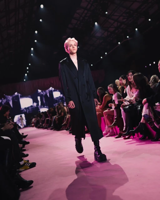 A fashion runway featuring a person with light-pink hair and a dark coat, walking towards the camera with an audience in the background.