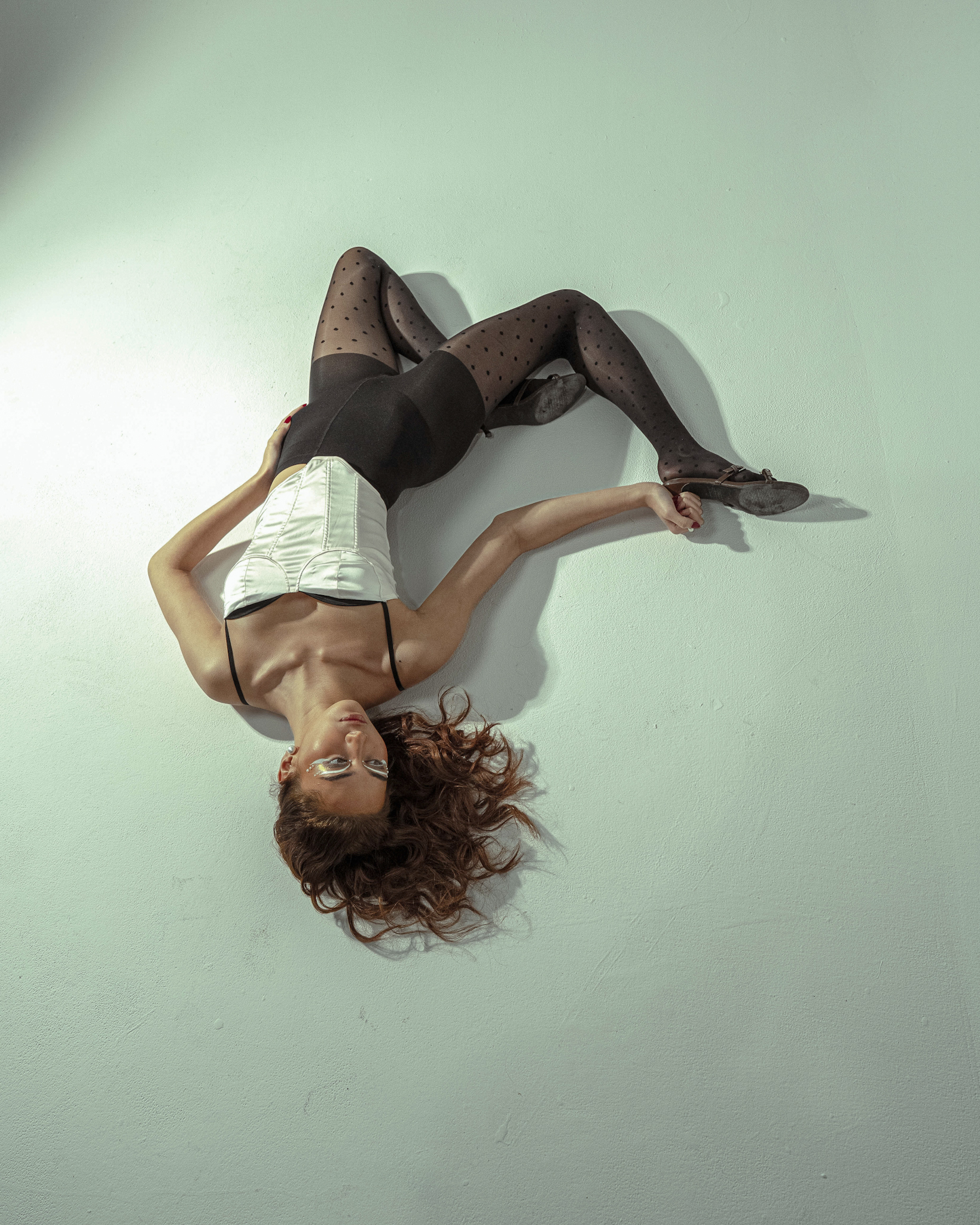 Model lying on a light grey studio floor wearing a white corset, black briefs, and dotted sheer tights.
