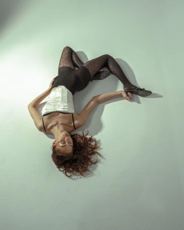 Model lying on a light grey studio floor wearing a white corset, black briefs, and dotted sheer tights.