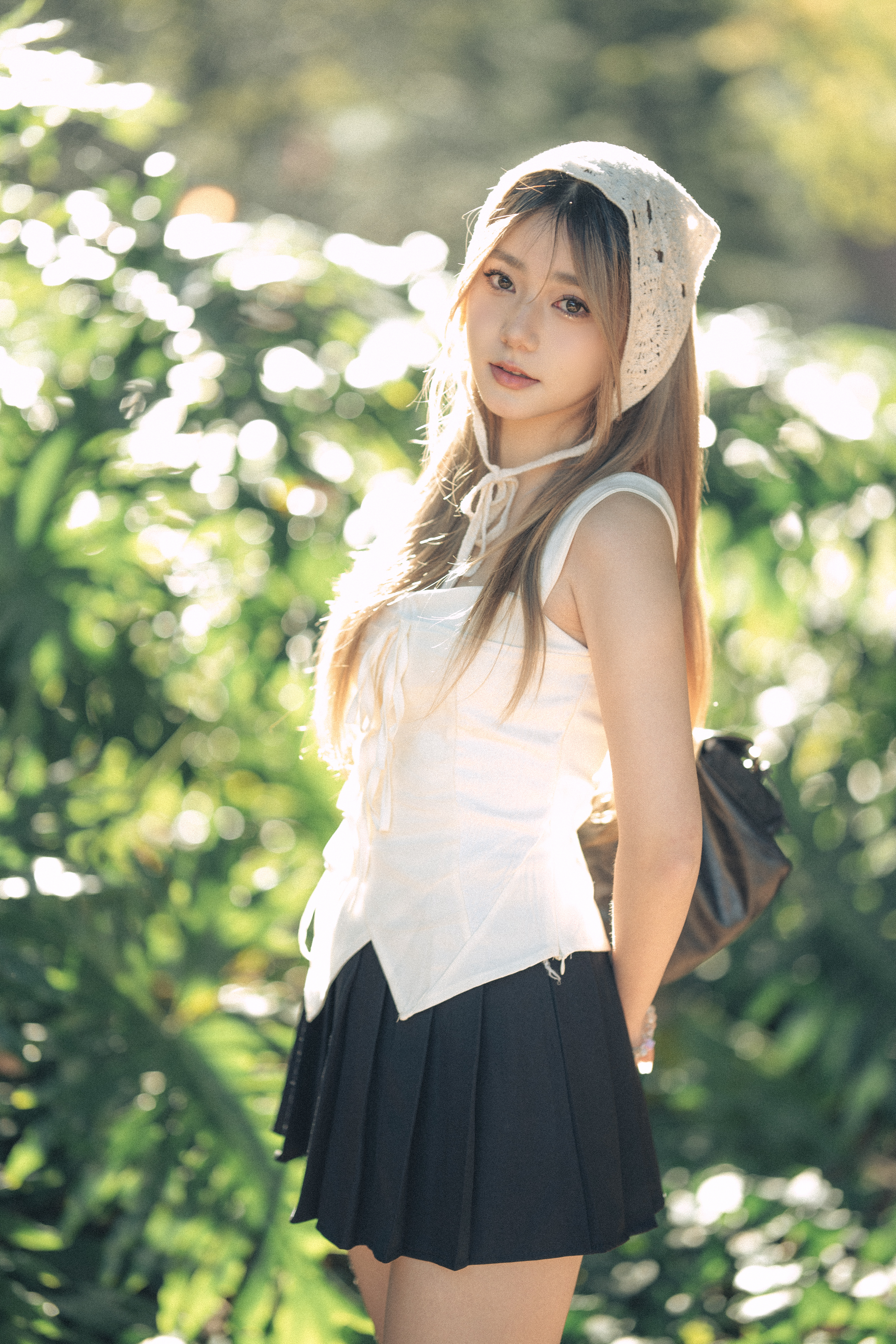 Young woman with long blonde hair and a white top stands outdoors in natural light with bokeh background, wearing a light headscarf.