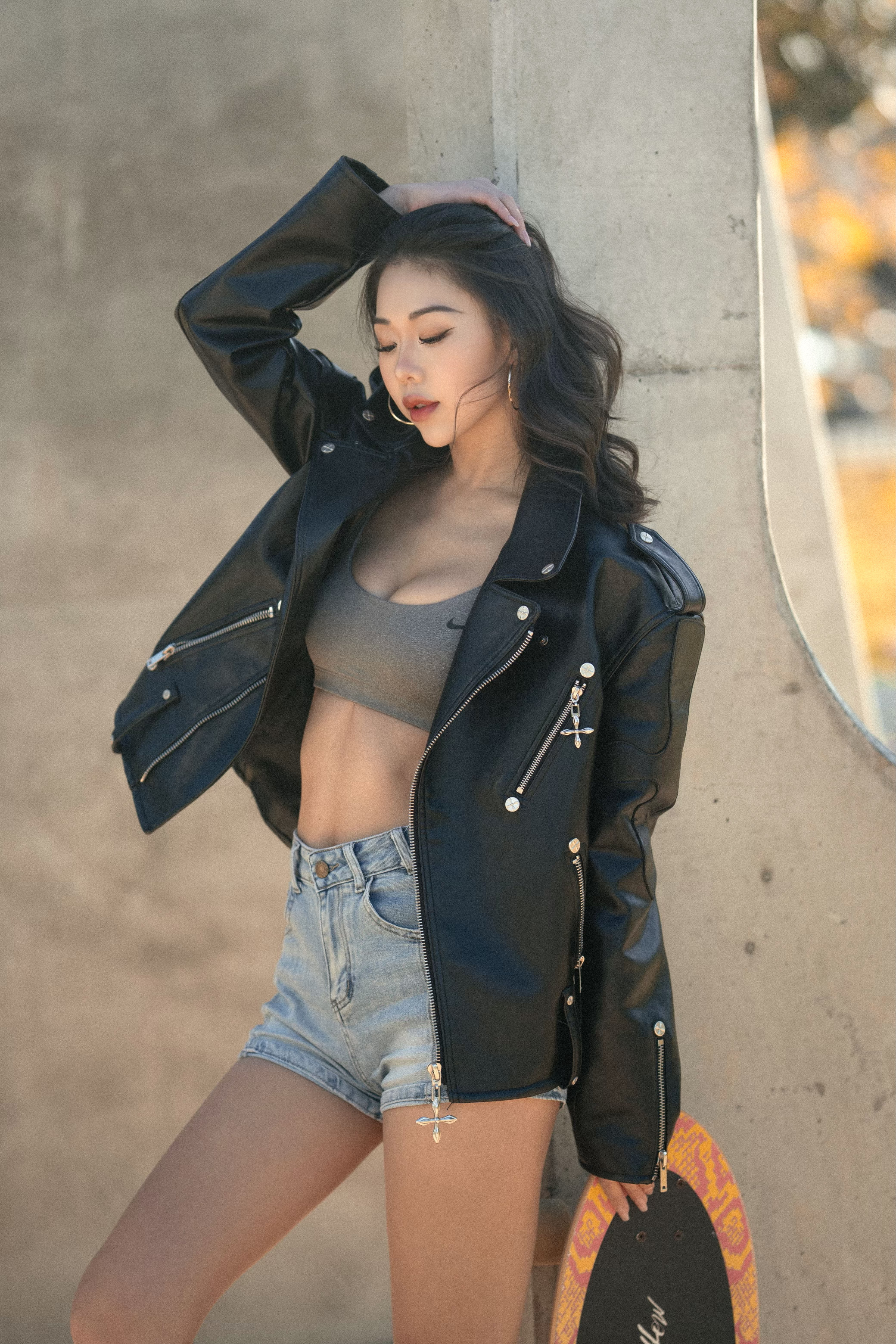 An Asian woman in a black leather jacket poses against a concrete wall, holding a skateboard, with soft lighting highlighting her features.