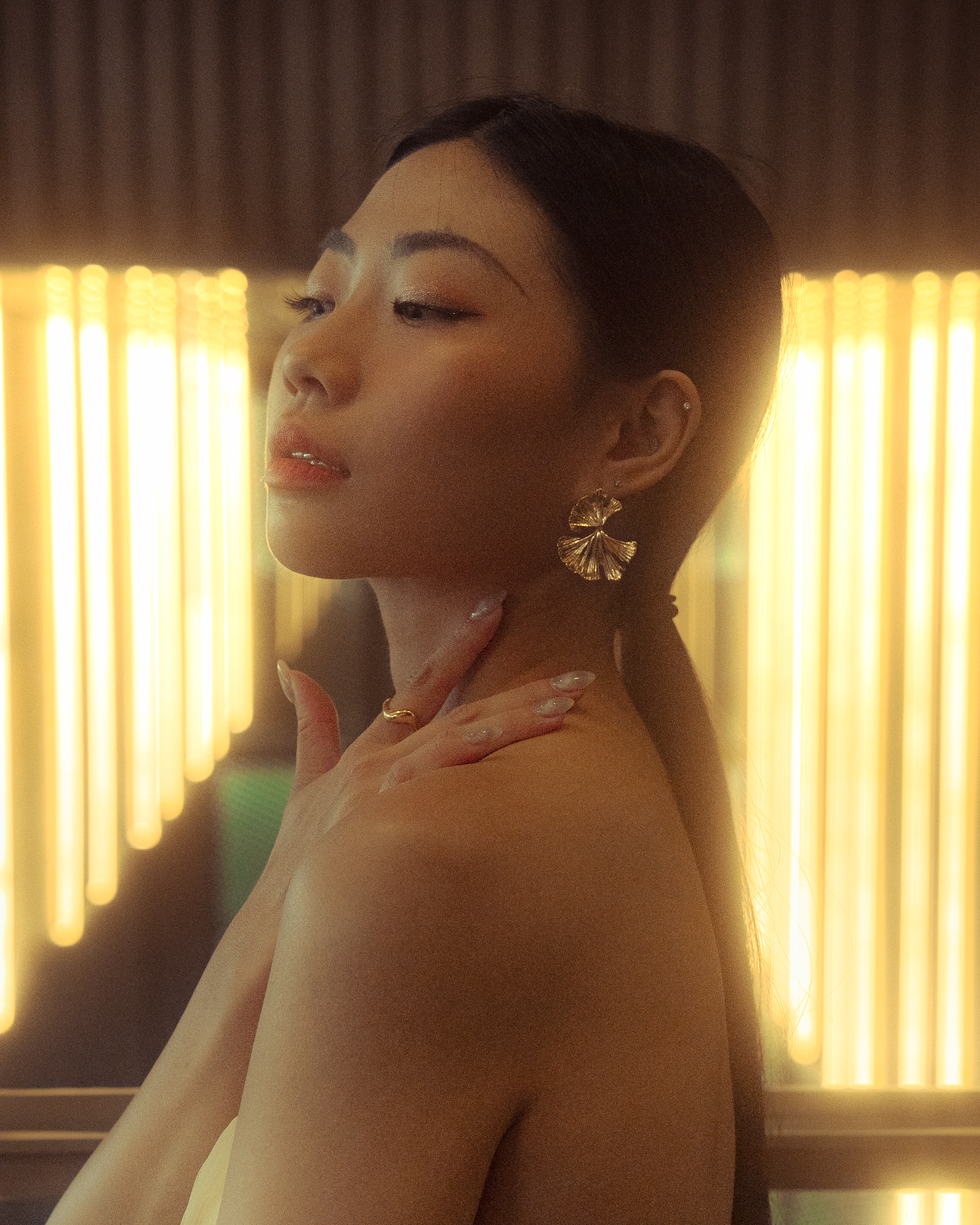 Close-up portrait of a young Asian woman with gold flower earrings and a bare shoulder posing indoors with a warm-toned, glowing background.