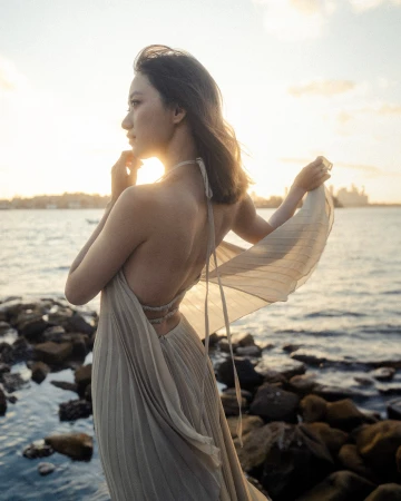 A woman in a cream halter neck pleated dress standing by rocky waterfront at sunset captured by Kavan Lui