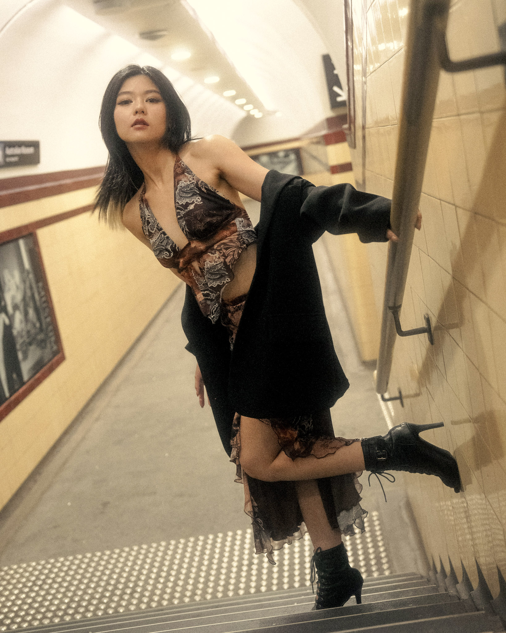 Stylish Asian woman poses on tiled stairs in a halter top, dress, and blazer in a brightly lit subway station with black ankle boots.
