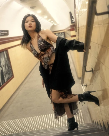 Woman in patterned halter top and dark coat posing on tiled subway station stairs with hand on rail.