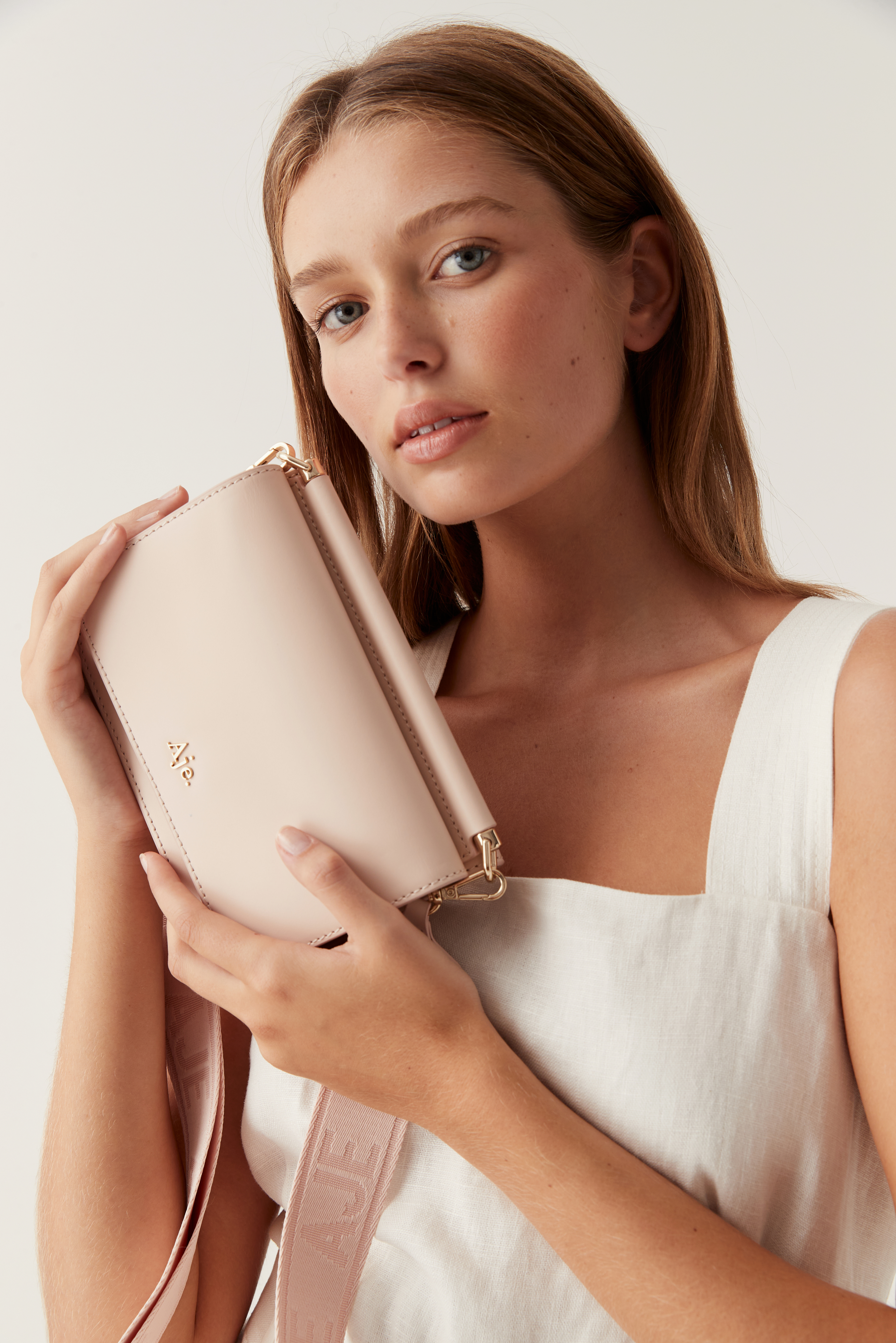 Woman holding a light pink purse with a strap and a logo detail on a white background in a studio setting.