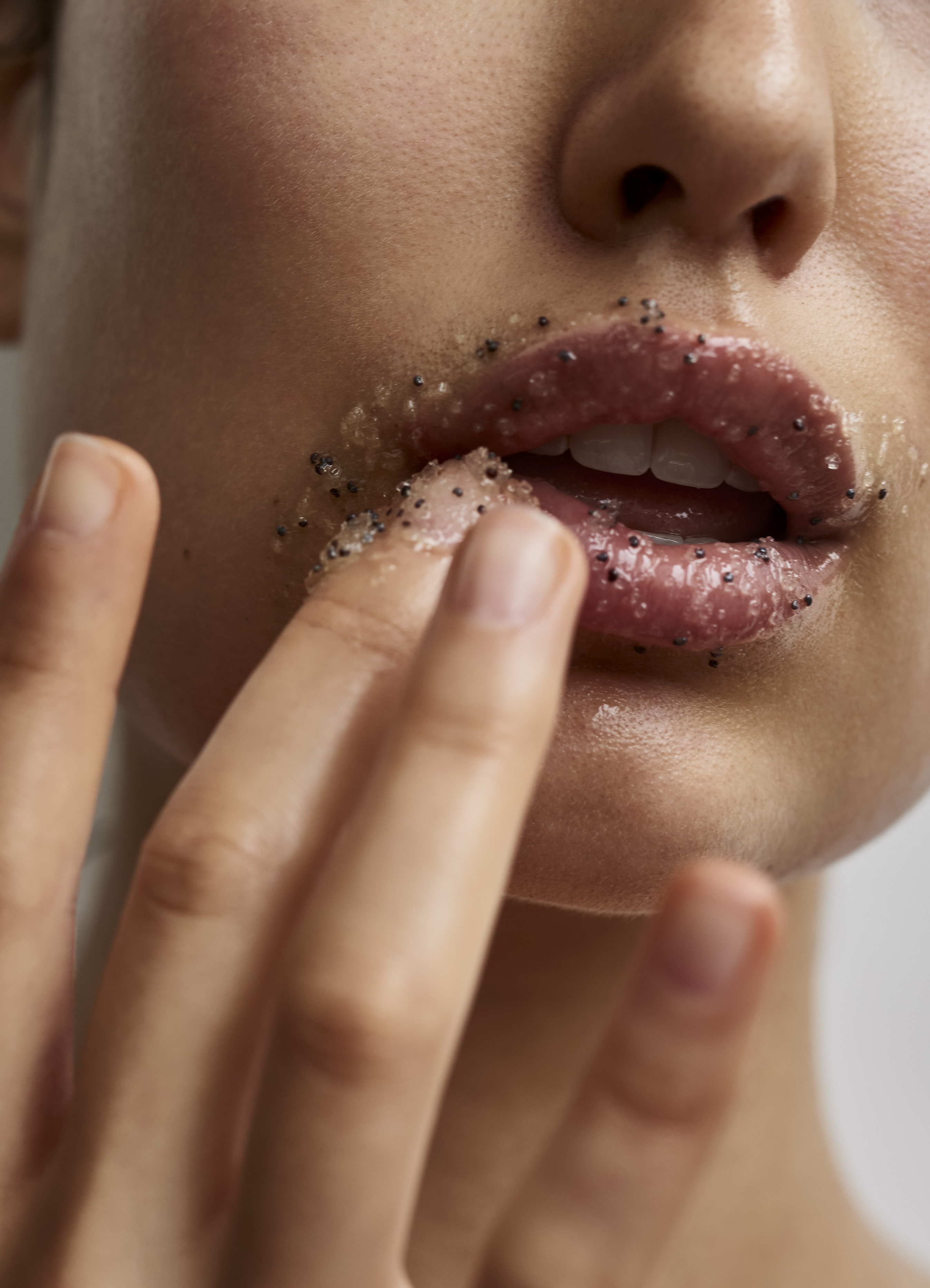 Close-up of a woman's face applying lip scrub to her lips with her finger, skincare and beauty concept, neutral background, studio lighting.