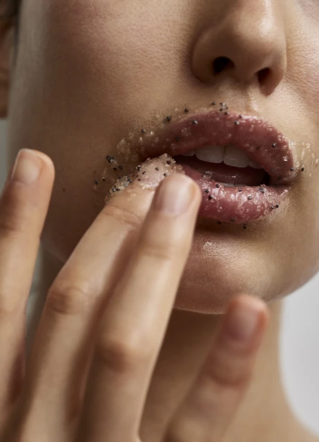 Close-up of a woman's face applying lip scrub to her lips with her finger, skincare and beauty concept, neutral background, studio lighting.
