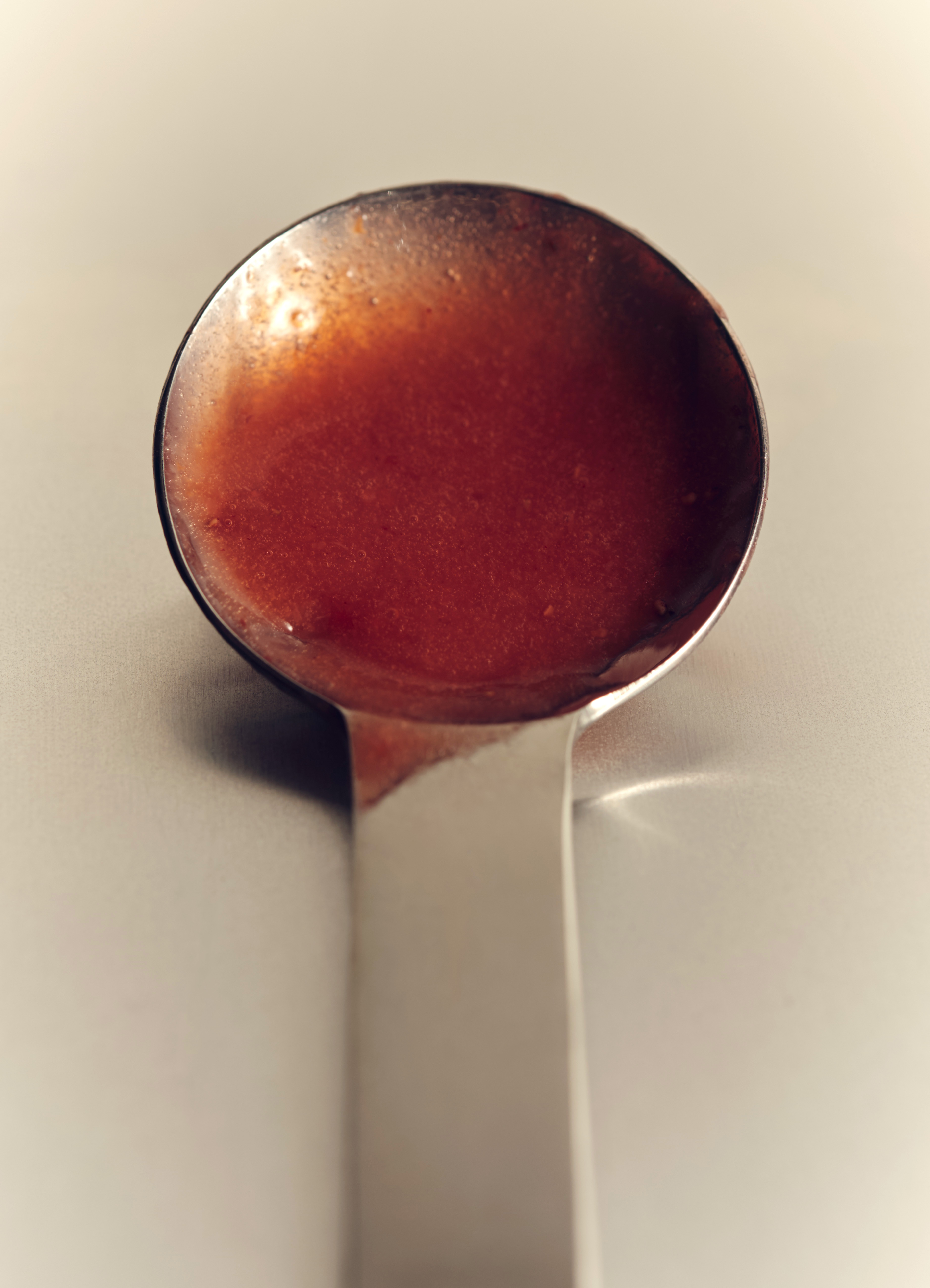 Overhead close-up of a silver metal ladle filled with a bright red sauce or soup on a white surface, casting soft shadows.