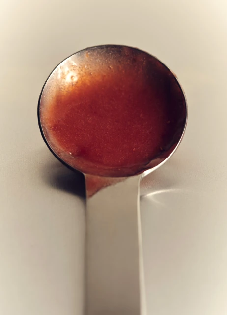 Overhead close-up of a silver metal ladle filled with a bright red sauce or soup on a white surface, casting soft shadows.