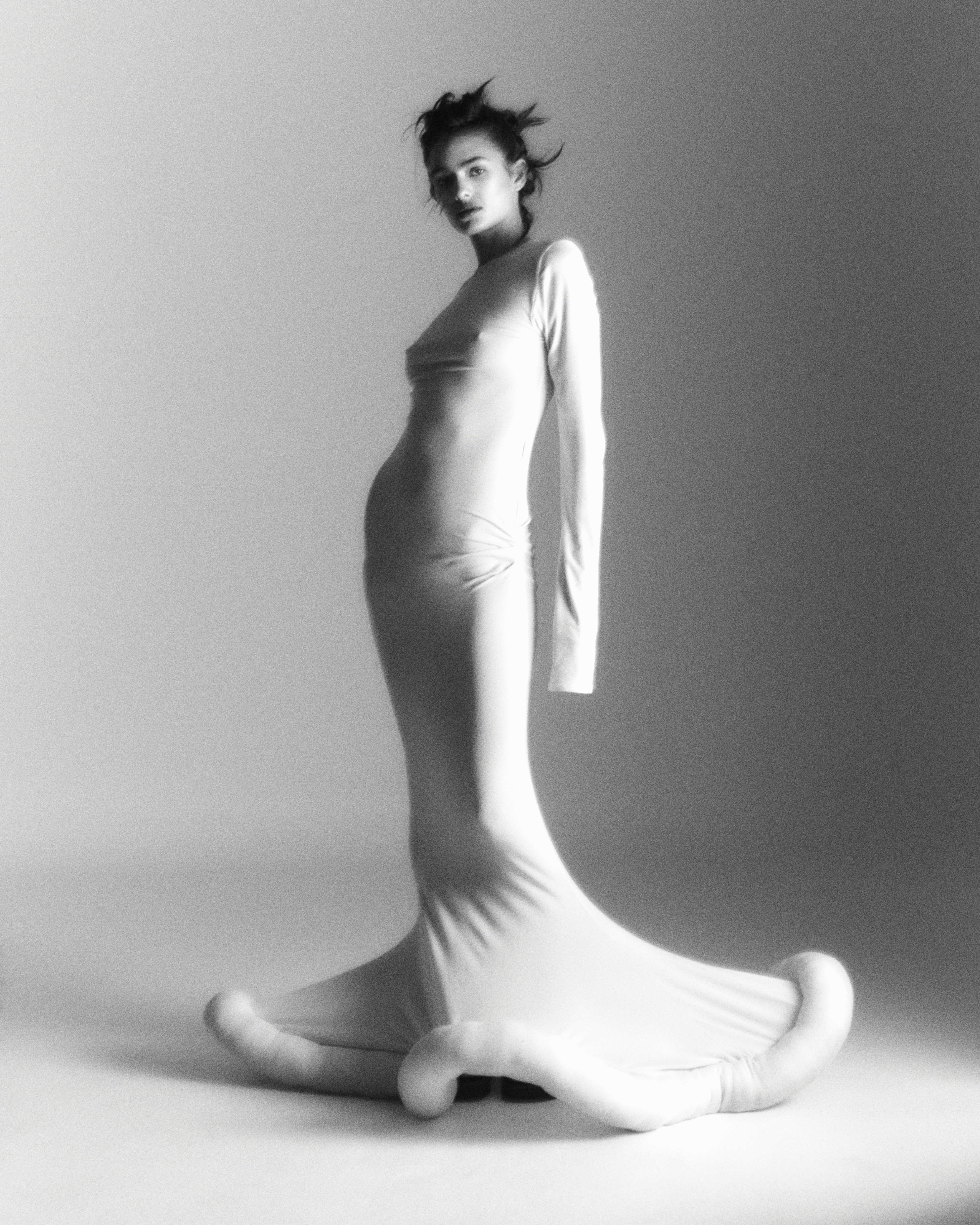 Monochrome studio shot of a woman in a long-sleeve dress with unique extended hem, striking a pose in front of a light grey background.