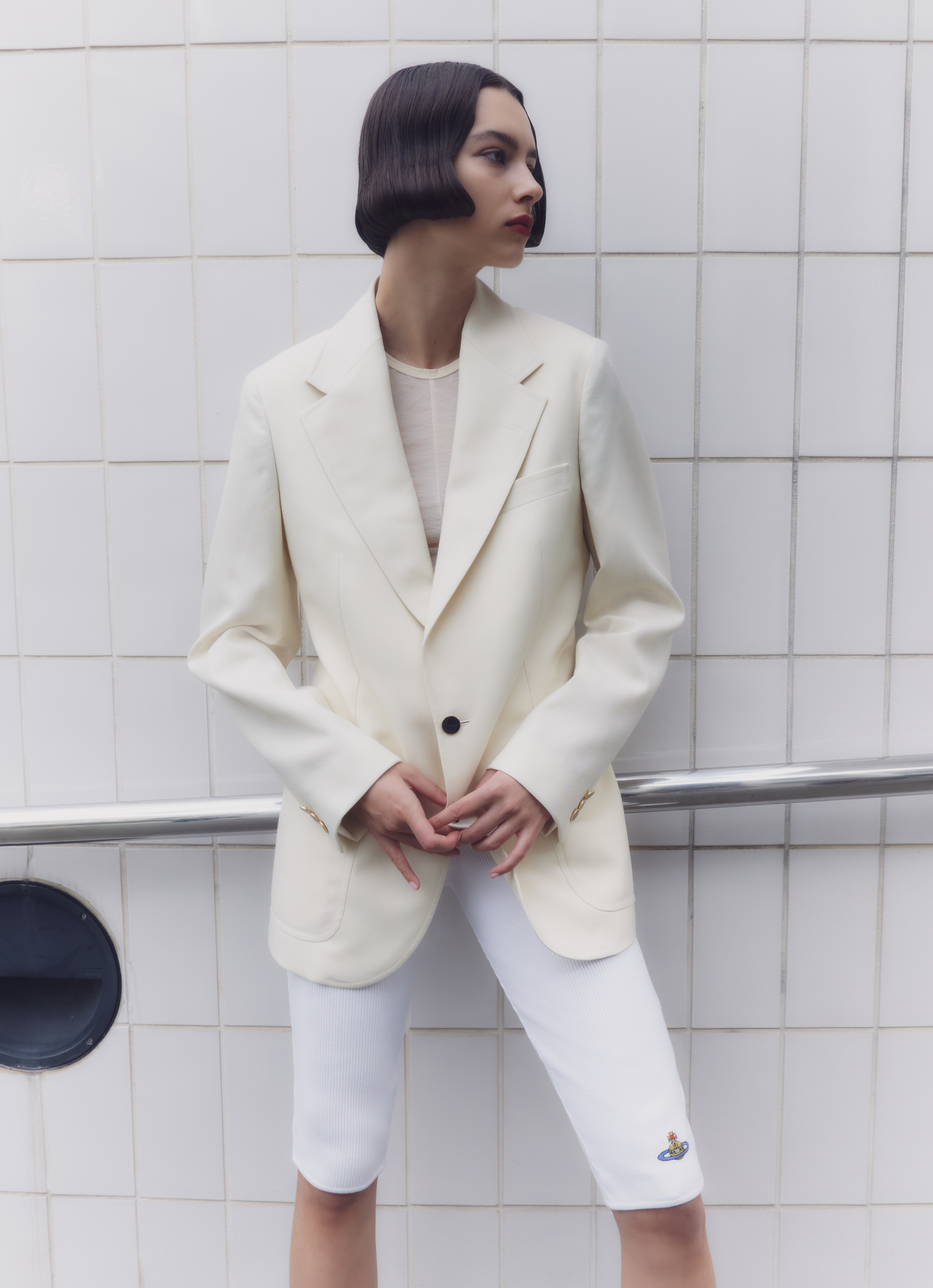 A woman poses in a cream blazer and white shorts against a white tiled wall, showcasing a fashion-forward and modern style.