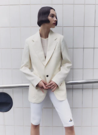 A woman poses in a cream blazer and white shorts against a white tiled wall, showcasing a fashion-forward and modern style.