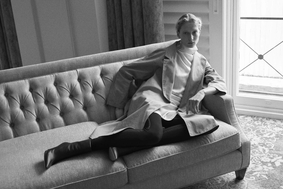 An adult female poses on a grey upholstered couch, wearing a white top, dark leggings, a coat, and dark pointed-toe boots, in a room with neutral tones.