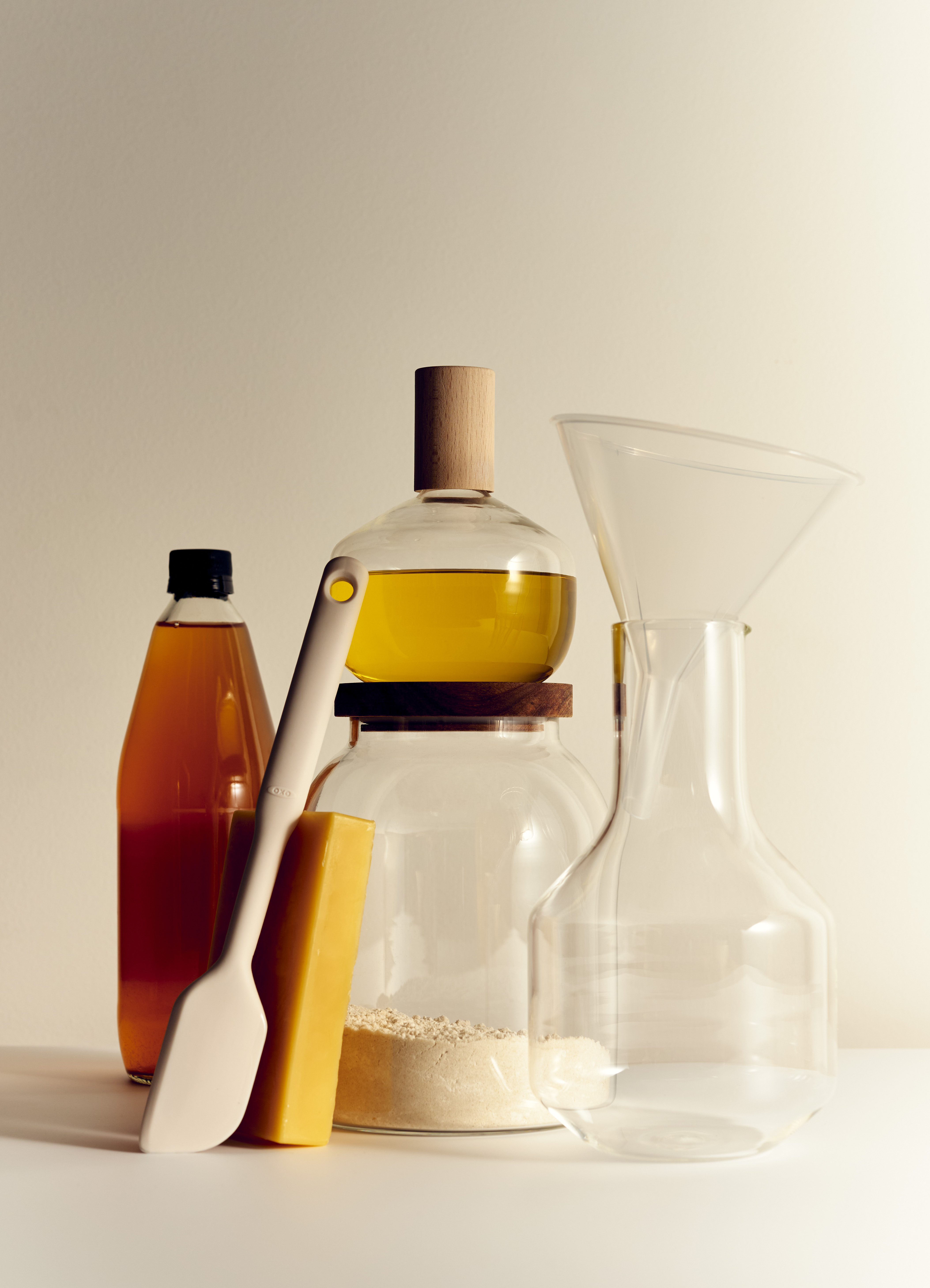 Still life of kitchenware and ingredients including oil, vinegar, spatula, and funnel, against a light beige background in soft lighting.