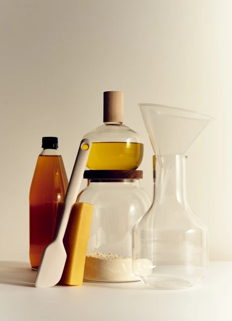 Still life of kitchenware and ingredients including oil, vinegar, spatula, and funnel, against a light beige background in soft lighting.