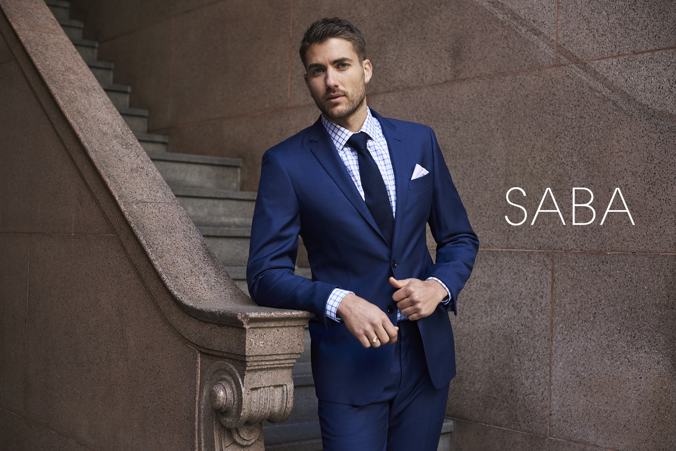 Fashion shoot of a man in a blue suit leaning against a marble staircase with a neutral stone wall, showcasing a stylish and professional look.