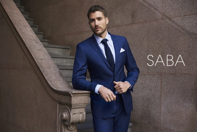 Fashion shoot of a man in a blue suit leaning against a marble staircase with a neutral stone wall, showcasing a stylish and professional look.