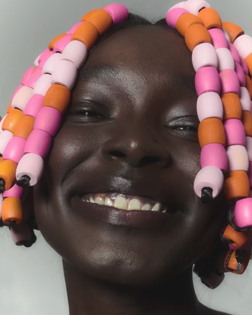Close-up portrait of a smiling Black woman with beaded hair accessories in pink and orange against a neutral-toned background.