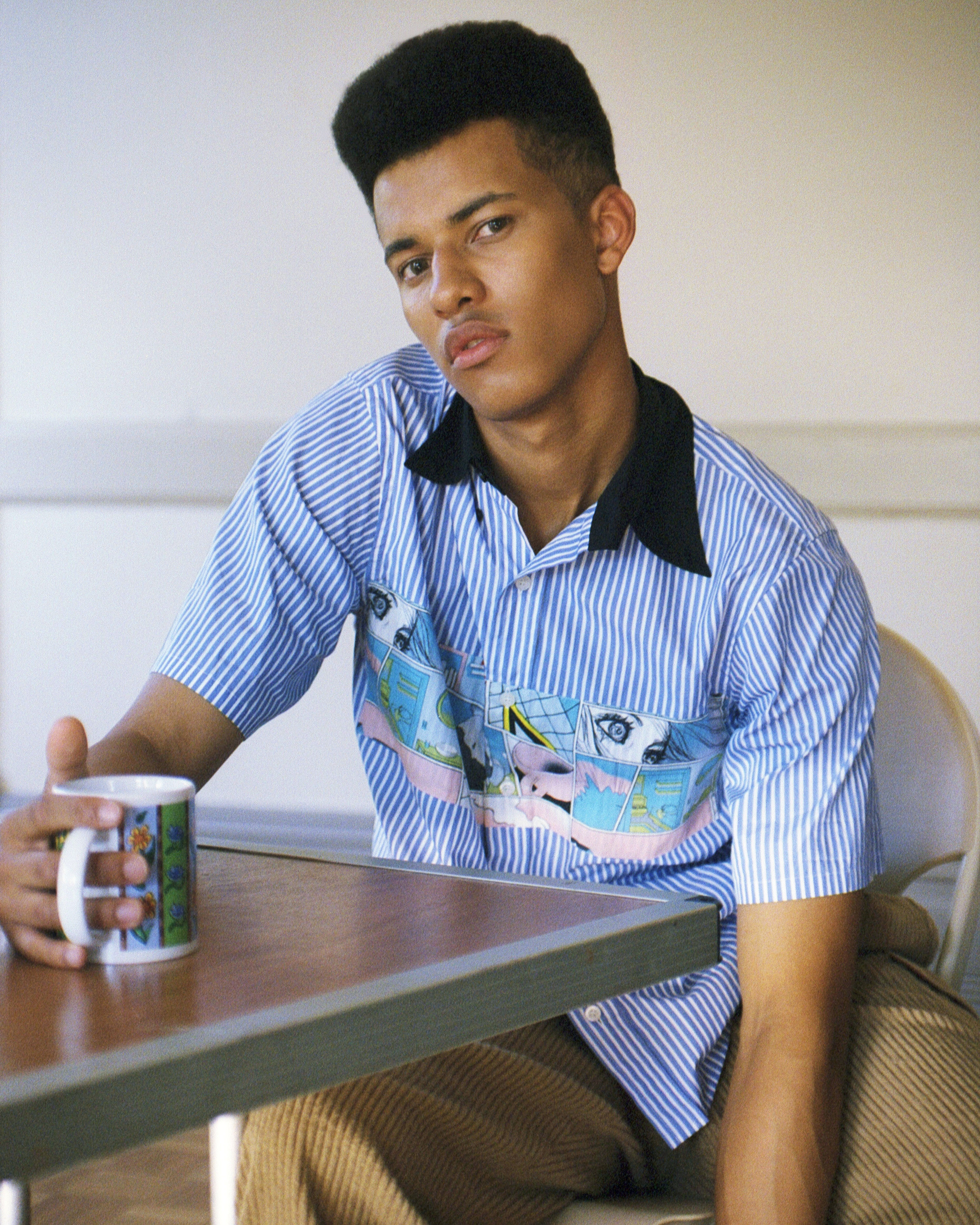 Young man in a striped blue short sleeve shirt seated at a wooden table holding a patterned mug
