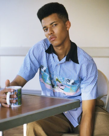 Young man in a striped blue short sleeve shirt seated at a wooden table holding a patterned mug